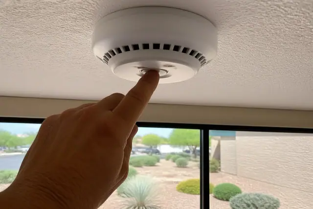 A hand pressing the test button on a ceiling-mounted smoke detector in Phoenix, AZ.