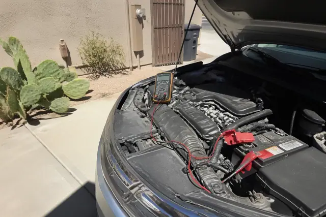 Multimeter connected to a vehicle battery for alternator and starter diagnostics on a residential concrete pad in Phoenix