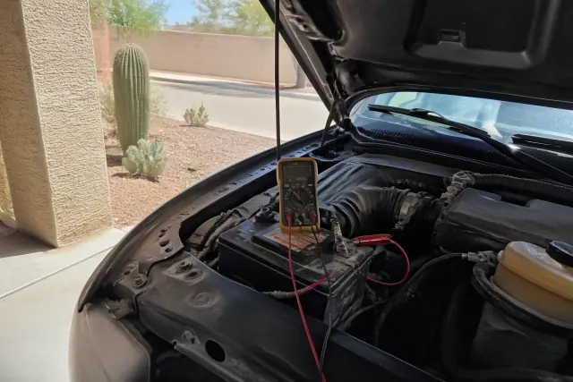Technician testing a car battery with a multimeter under a residential carport in Phoenix, AZ.
