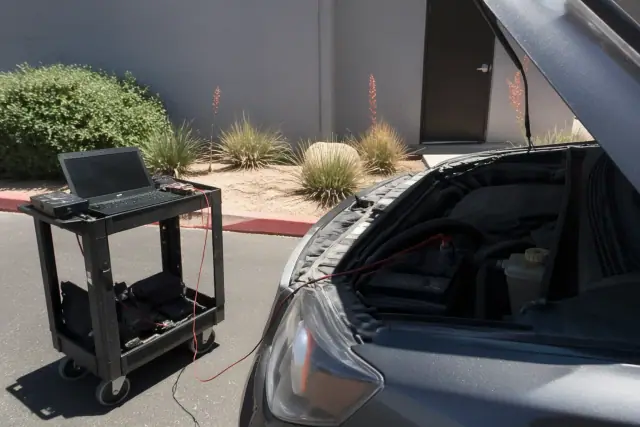 Technician connecting diagnostic cables to a vehicle engine in a paved utility area in Phoenix, AZ.