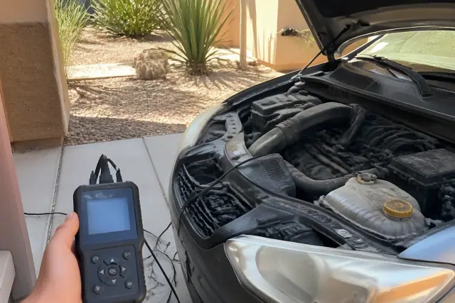 A mechanic using a handheld diagnostic scanner on a car engine in a residential driveway in Phoenix, AZ.