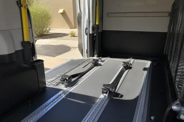 Wheelchair tie-down straps anchored in a transport vehicle in Phoenix, AZ, with desert landscaping visible outside.