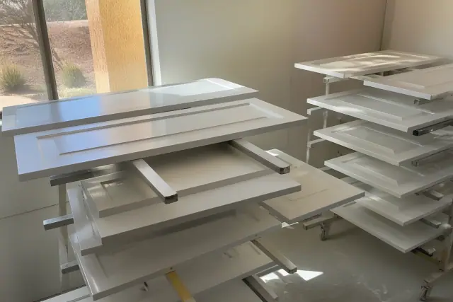 Cabinet doors drying on racks in a workspace in Phoenix, AZ, visible through a window with desert stucco walls.