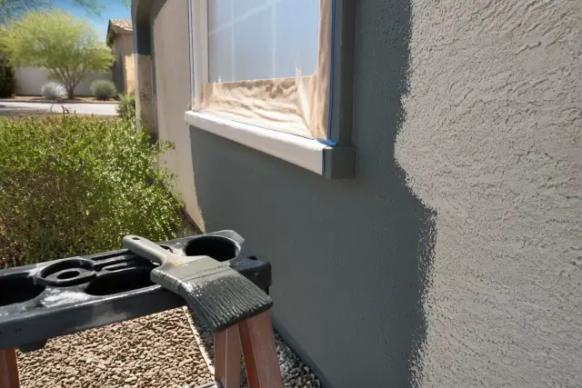 A paint brush on a ladder tray against a partially masked stucco wall at an exterior painting job in Peoria, AZ.