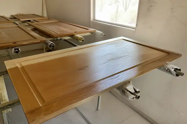 Cabinet door being stained and topcoated in a Phoenix, AZ workshop with sunlight and stucco walls.