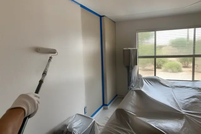 A technician paints a wall in a Phoenix, AZ residential living room with furniture covered in plastic sheeting.