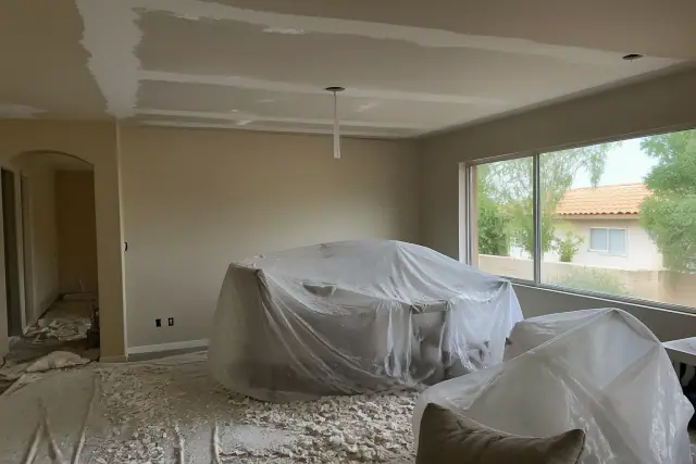 Popcorn ceiling removal in progress in a Scottsdale, AZ home with furniture covered in plastic and debris on the floor.