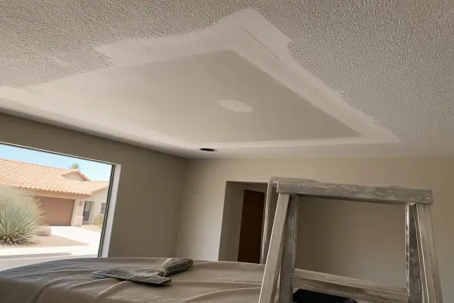 Sanded drywall patch on a ceiling in Phoenix, AZ, with a ladder and joint knife on a drop cloth in a home.
