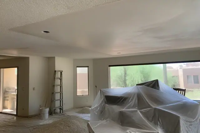 A residential room in Phoenix, AZ, with furniture covered in plastic and popcorn ceiling debris on drop cloths.