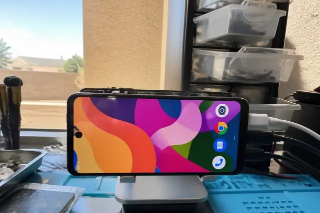 A smartphone screen being tested with diagnostic cables on a workbench in Phoenix, AZ, with sunlight on a stucco wall.