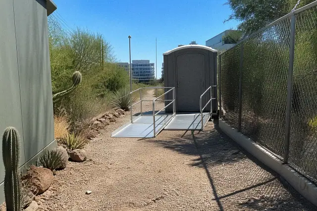 ADA-compliant portable toilet with ramp on a gravel path in Phoenix, AZ, surrounded by desert landscaping and fencing.