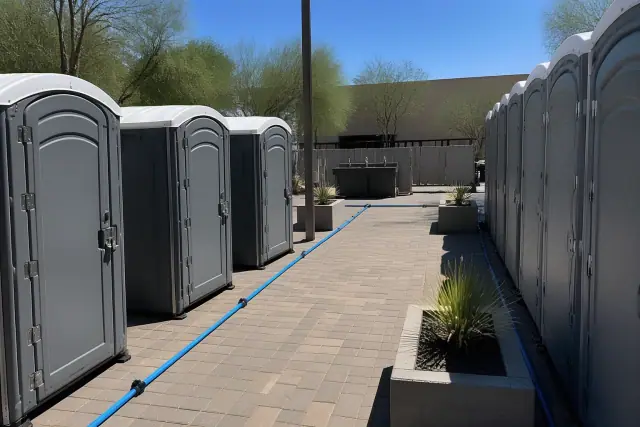 Portable toilets and sinks on a paved courtyard in Phoenix, AZ, with visible wastewater lines and desert landscaping.