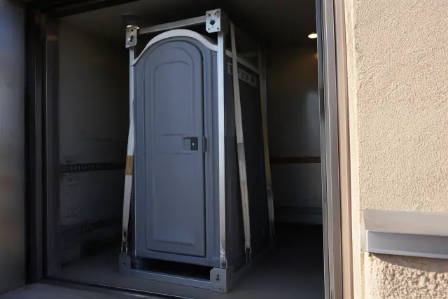 Narrow-profile portable restroom unit with hoisting straps inside an industrial lift bay in Peoria, AZ.