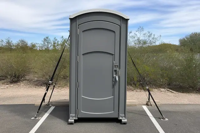 Grey portable toilet secured with stabilization straps on an asphalt parking lot in Peoria, AZ.