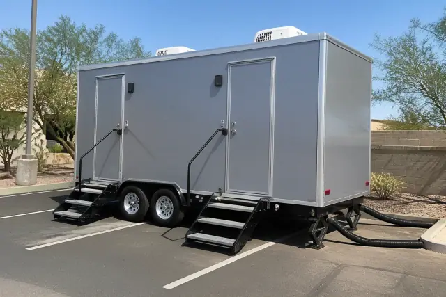 Luxury restroom trailer parked on an asphalt lot in Mesa, AZ, showing drainage hoses and desert landscaping.
