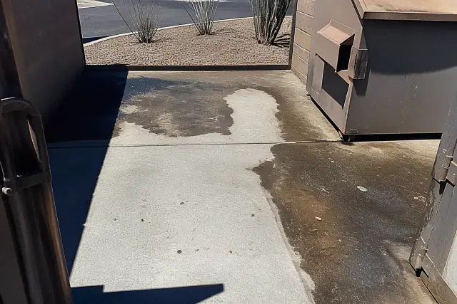 Cleaned concrete dumpster pad showing grease removal next to metal trash bins in Goodyear, AZ.