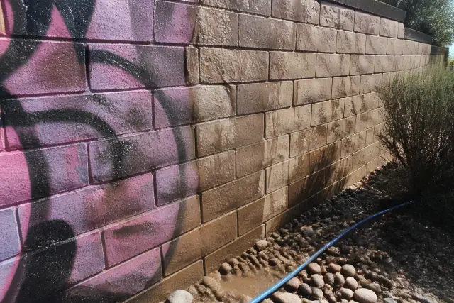 A masonry wall in Buckeye, AZ showing the progress of graffiti removal from brick, with desert landscaping in the background.