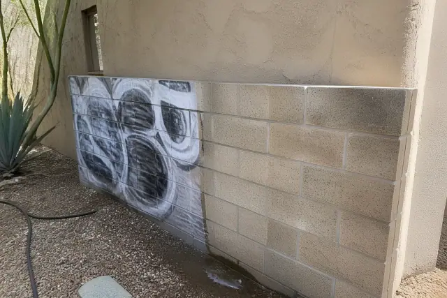 A partially cleaned brick wall showing the removal of graffiti in Phoenix, AZ, with desert landscaping in the background.