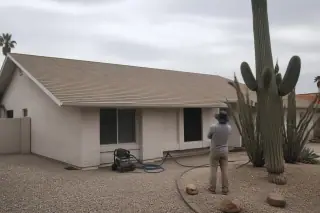 Contractor inspecting a clean asphalt-shingle roof and coiled wash hose at a stucco home in Phoenix, AZ.
