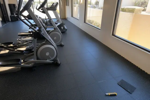 Installer fitting rubber floor tiles in a commercial gym in Phoenix, AZ, with cardio machines and tools nearby.