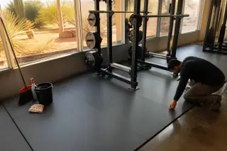 Worker inspecting rubber floor mats under a squat rack in a Phoenix, AZ gym with desert sunlight.