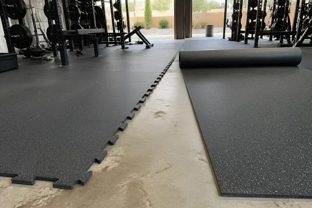 Technician installing rubber gym tiles on a concrete slab in a Phoenix, AZ facility with rolled flooring nearby.
