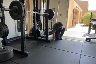 Worker checking a rubber floor seam under a squat rack in a Phoenix, AZ gym with bright desert light.