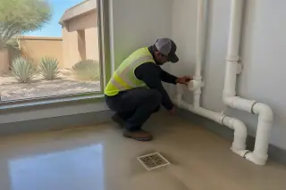Technician in a high-visibility vest inspecting PVC drainage pipes in a Phoenix, AZ utility room.