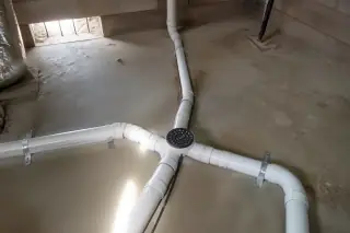 Cleaned PVC drainage pipes and a central floor drain in a Phoenix, AZ utility room with dry soil near the foundation.