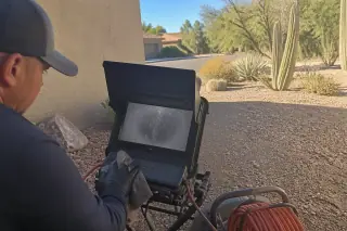 Technician wiping down a sewer camera monitor in a Phoenix, AZ yard with desert landscaping in the background.