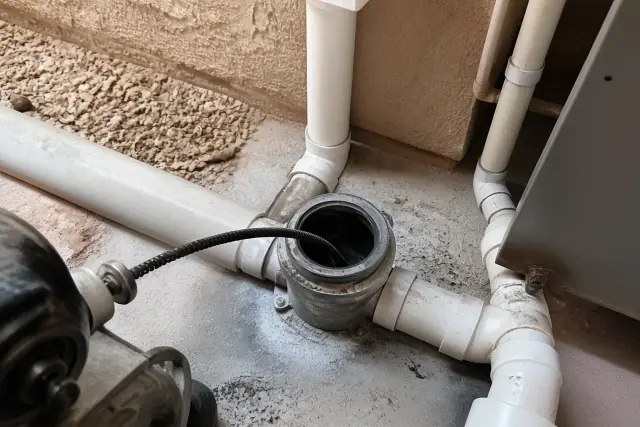 Motorized cable augering in a Phoenix, AZ basement utility room with a steel cable inserted into PVC drainage pipes.