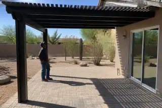 Worker inspecting a completed wood ramada over a stone patio in a Phoenix, AZ backyard with desert landscaping.