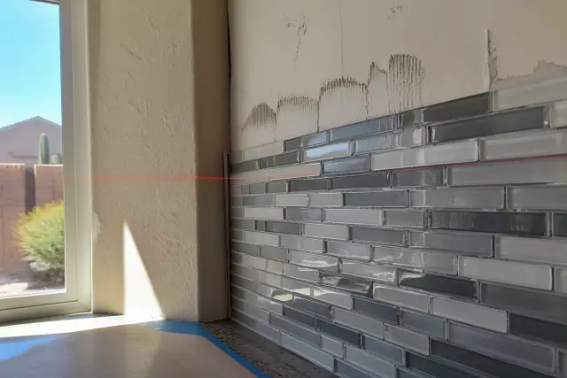 A red laser level beam across a partially installed glass and mosaic tile kitchen backsplash in Phoenix, AZ.