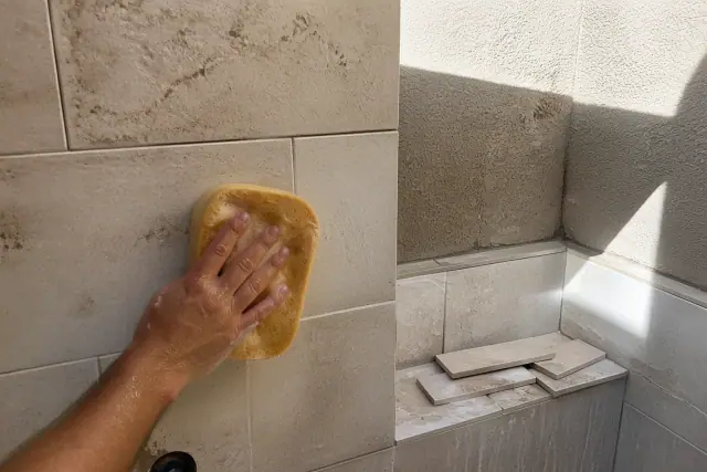 A hand wiping grout from new stone shower tiles in a Phoenix, AZ home under bright desert sunlight.
