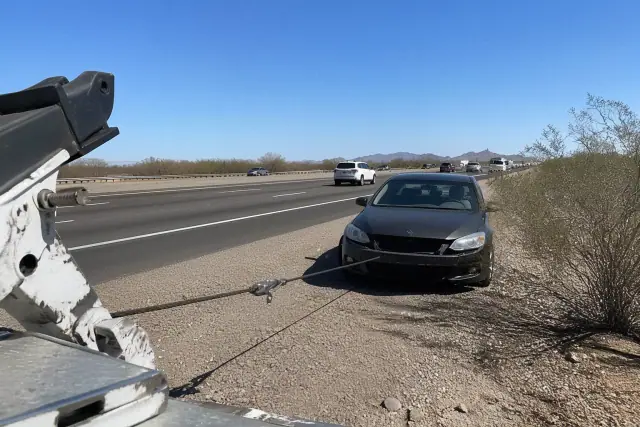A disabled vehicle being recovered by a winch on a gravel highway shoulder in Phoenix, AZ.