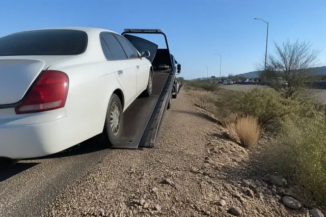 Flatbed tow truck loading a disabled vehicle on a roadside in Phoenix, AZ, under bright desert sunlight.