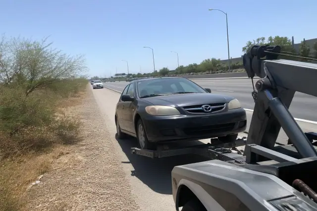A local towing truck hooked to a disabled car on a highway shoulder in Phoenix, AZ.
