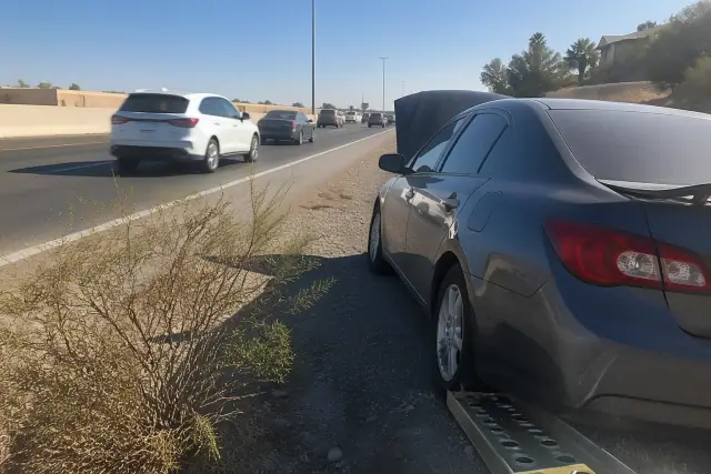 A disabled vehicle parked on the gravel shoulder of a highway in Mesa, AZ with desert scrub in the background.