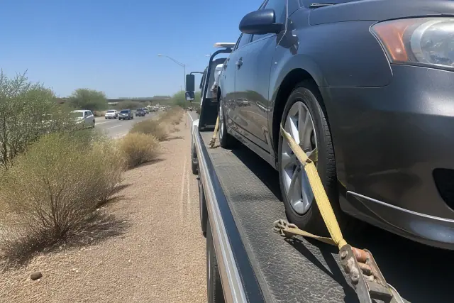 A disabled vehicle secured with yellow straps to a flatbed tow truck on a gravel shoulder in Scottsdale, AZ.