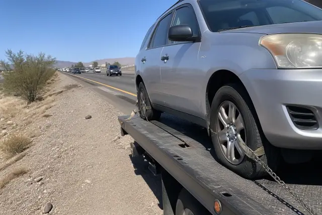 A disabled vehicle secured by chains and straps onto a flatbed tow truck on a highway shoulder in Phoenix, AZ.