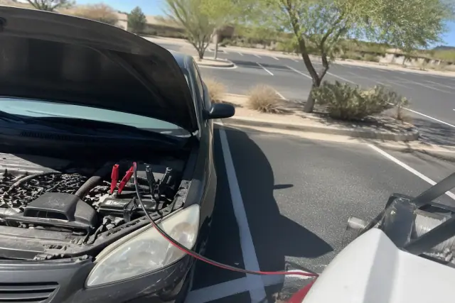 Technician using jumper cables on a car battery with an open hood in a Phoenix, AZ parking lot with desert landscaping.