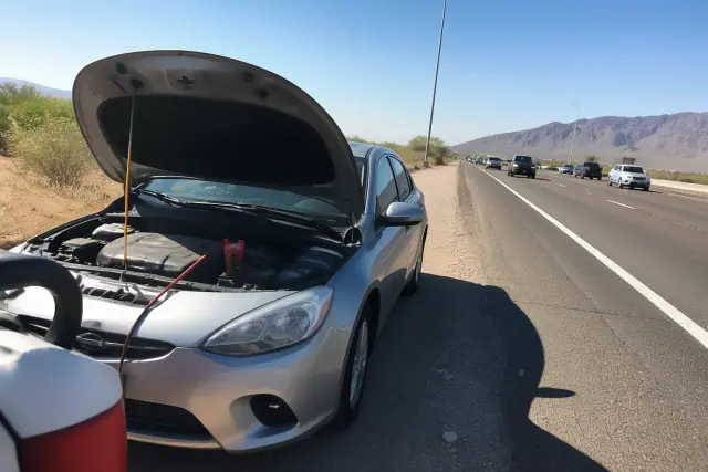 A disabled car with its hood open and jumper cables on a gravel highway shoulder in Goodyear, AZ.