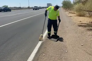 Technician using a broom and dustpan to clean road debris on a highway shoulder in Phoenix, AZ.