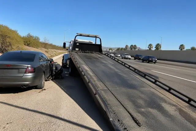 A tow truck winching a disabled vehicle onto its hydraulic bed on a highway shoulder in Phoenix, AZ.