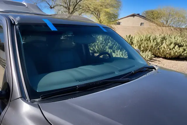 Newly installed windshield with retention tape on a vehicle at a work site in Tempe, AZ, with prickly pear cacti nearby.