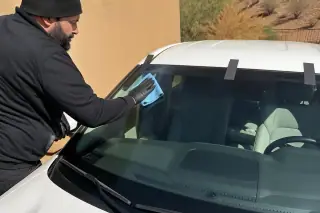 Technician cleaning a newly installed windshield with retention tape on a car in a sunny Phoenix, AZ driveway.