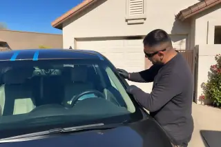 Technician inspecting a new car windshield with retention tape in a sunny residential driveway in Phoenix, AZ.