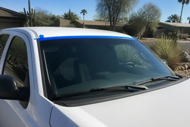 A newly installed windshield with retention tape on a car parked in a sunny, xeriscaped front yard in Phoenix, AZ.