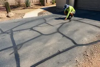 Technician inspecting black crack sealant on an asphalt driveway with desert landscaping in Scottsdale, AZ.