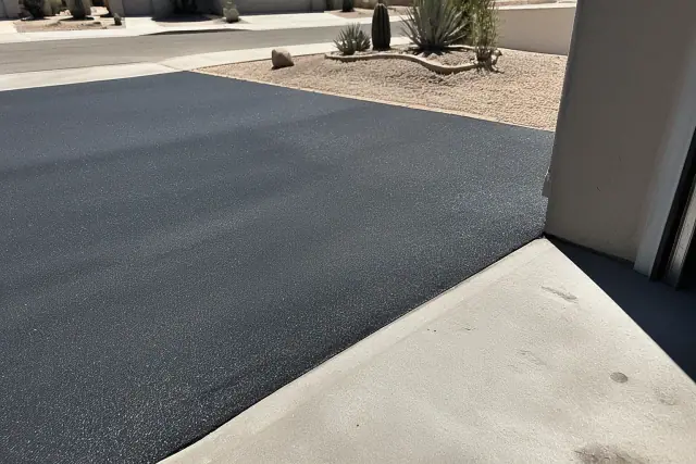 Newly sealed black asphalt driveway meeting a concrete garage slab in a residential yard in Tempe, AZ.